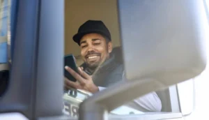 Smiling truck driver sitting in the cab and looking at his smartphone
