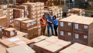 Three workers stand in a warehouse surrounded by stacks of large cardboard boxes
