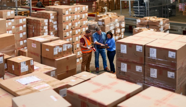 Three workers stand in a warehouse surrounded by stacks of large cardboard boxes