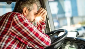 Frustrated truck driver in a red plaid shirt resting his head on the steering wheel inside the cab