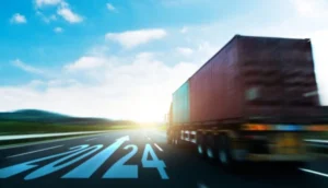 A large cargo truck drives on an open highway toward a bright horizon, with "2024" marked on the road