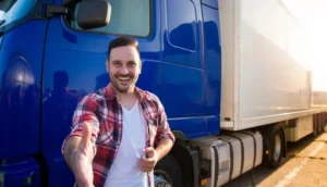 A smiling truck driver extends a handshake while standing in front of a blue semi-truck