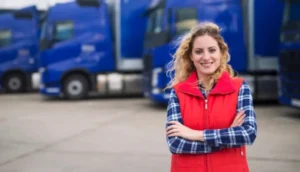 Woman in red vest and plaid shirt smiling confidently in front of a row of parked blue transport trucks